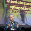 Mies puhuu lavalla. Taustalla näkyy tapahtuman mainoskuva, jossa lukee Missiologinen symposium.