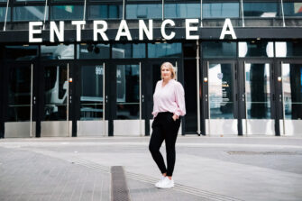 Kuvassa on Johanna Vauto, jolla on vaalea polkkatukka, mustat, kapeat housut, valkoiset lenkkarit ja vaaleanpunainen pitkähihainen paita. Hän seisoo Nokia Arenan sisäänkäynnin edessä harmaalla kivetyksellä.